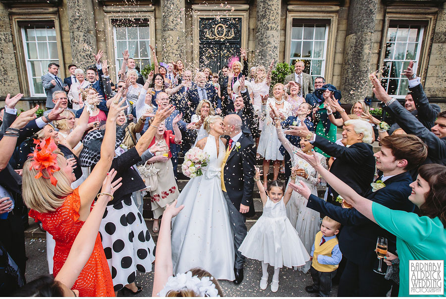 A bride and groom kiss beneath confetti surrounded by all their guests at a wedding venye called Denton Hall in Wharfedale near Ilkley in Yorkshire