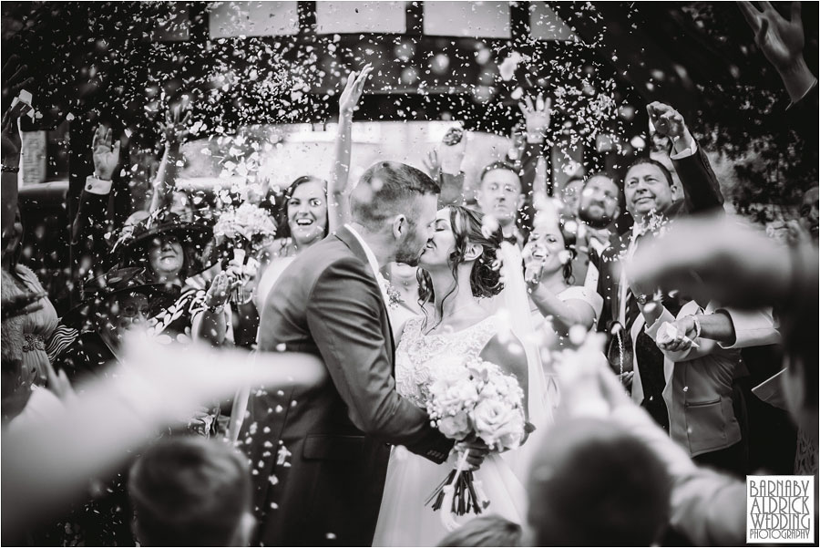A black and white photo of a bride and groom kissing as guests confetti at their wedding at Wood Hall near Wetherby in North Yorkshire
