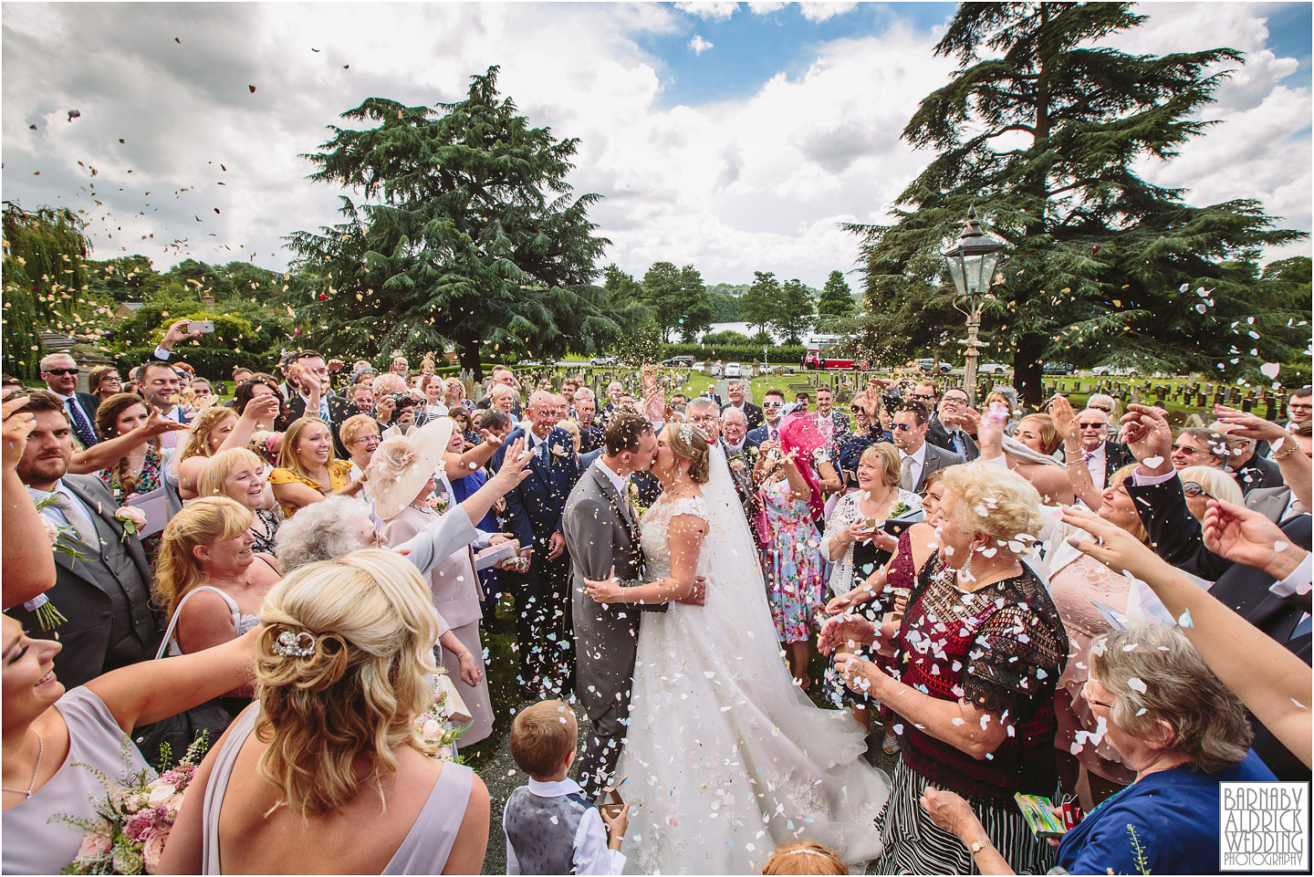A newly wed couple kiss under a dramatic confetti toss at their wedding at Alderford Lake in Shropshire