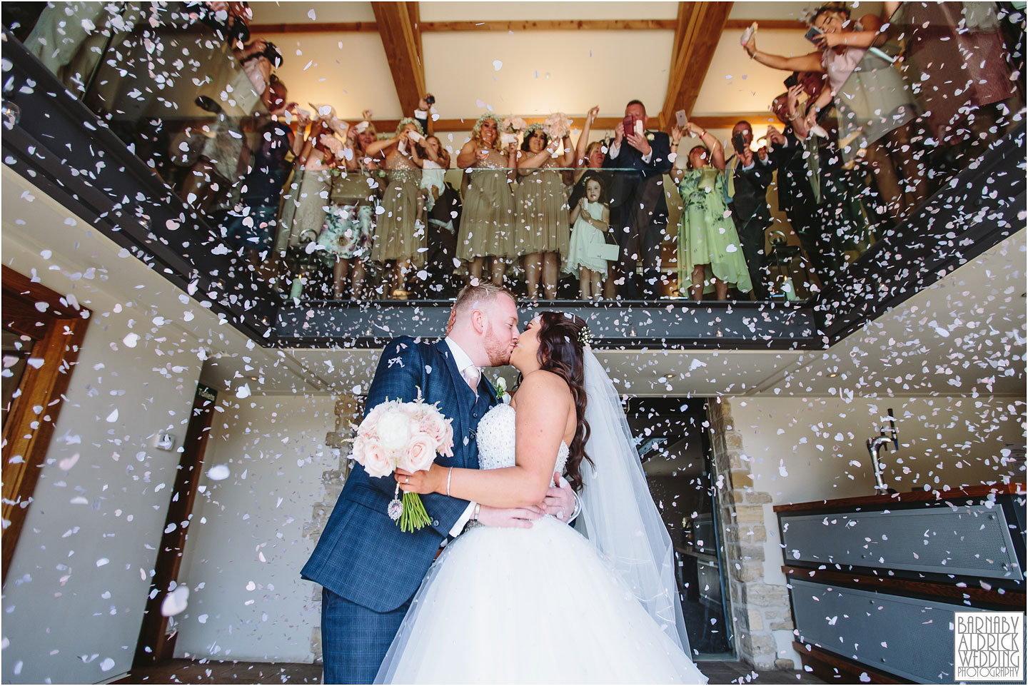 A bride and groom kiss under while guests throw confetti over them from the balcony during there beautiful barn ceremony at Priory Cottages wedding venue venue between Tadcaster and Wetherby