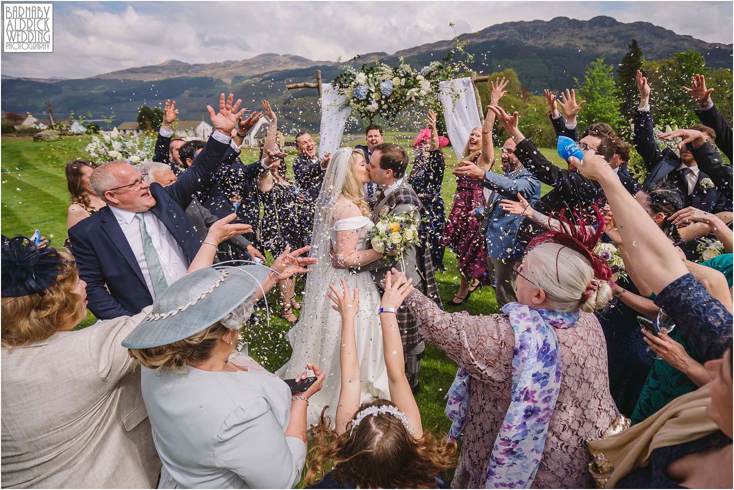A bride and groom in a kilt kiss under a dramatic confetti throw at their wedding at Carrick Castle Estate Lodge Barn on Loch Goil in Scotland